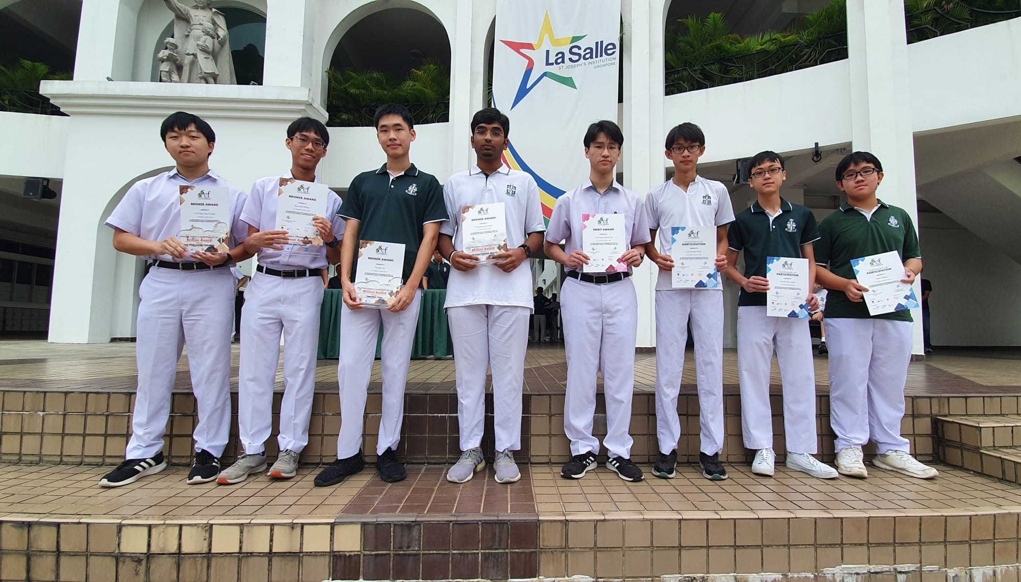 Eight male students in school uniforms hold award certificates outdoors in front of a La Salle banner.